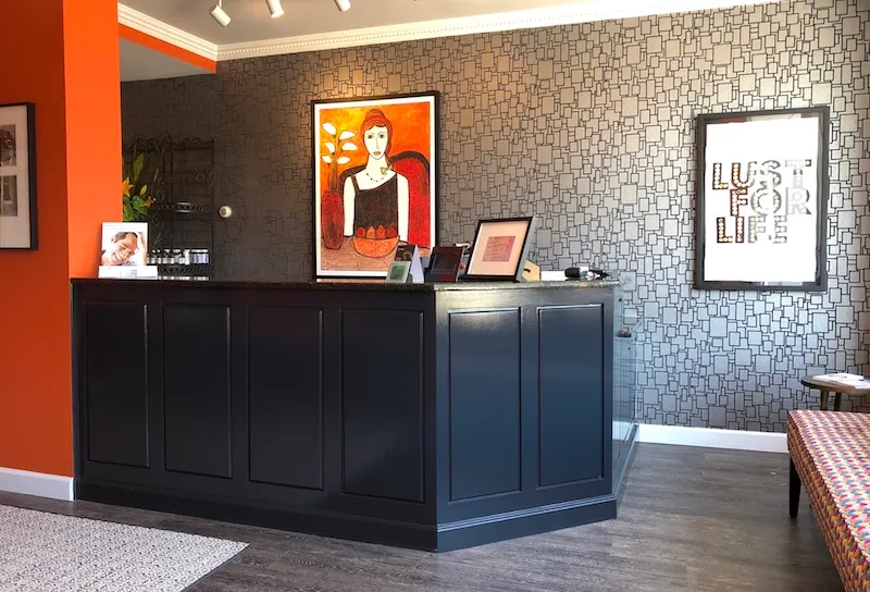 A modern dental reception area with a dark counter, patterned grey wallpaper, and vibrant orange accent walls featuring artistic portraits.