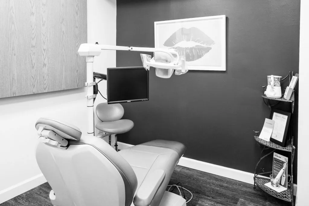 A dental treatment room with a minimalist aesthetic, featuring a grey patient chair against a dark grey wall with lip-themed artwork.
