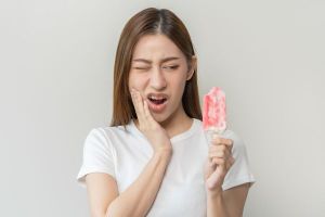 Young woman reacting to eating a cold popsicle with a hurt facial expression and touching her cheek to express tooth pain and sensitivity.
